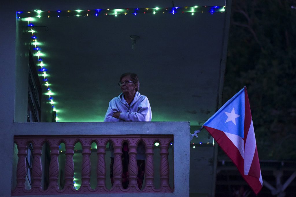 En esta foto del 21 de diciembre de 2017, Inocencia Rivera, madre de Eduardo González, un hombre que se suicidó tres semanas después del paso del huracán María, mira el anochecer en su balcón rodeada por luces navideñas que funcionan con energía solar y una bandera puertorriqueña. Foto: Carlos Giusti / AP.