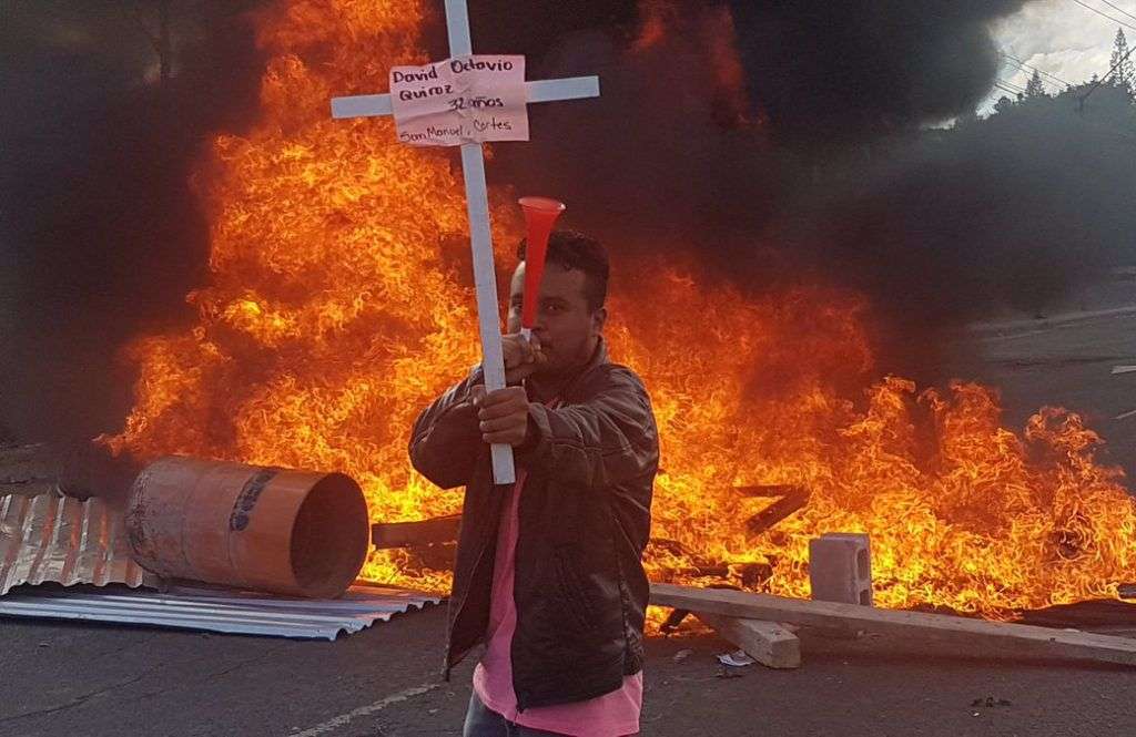 Manifestante muestra una cruz con el nombre de una de las víctimas de las represiones militares. Inicia la movilización de protesta contra toma de posesión de Juan Orlando Hernández. Foto: Twitter de Gilda Silvestrucci, Corresponsal en Honduras de Telesur.