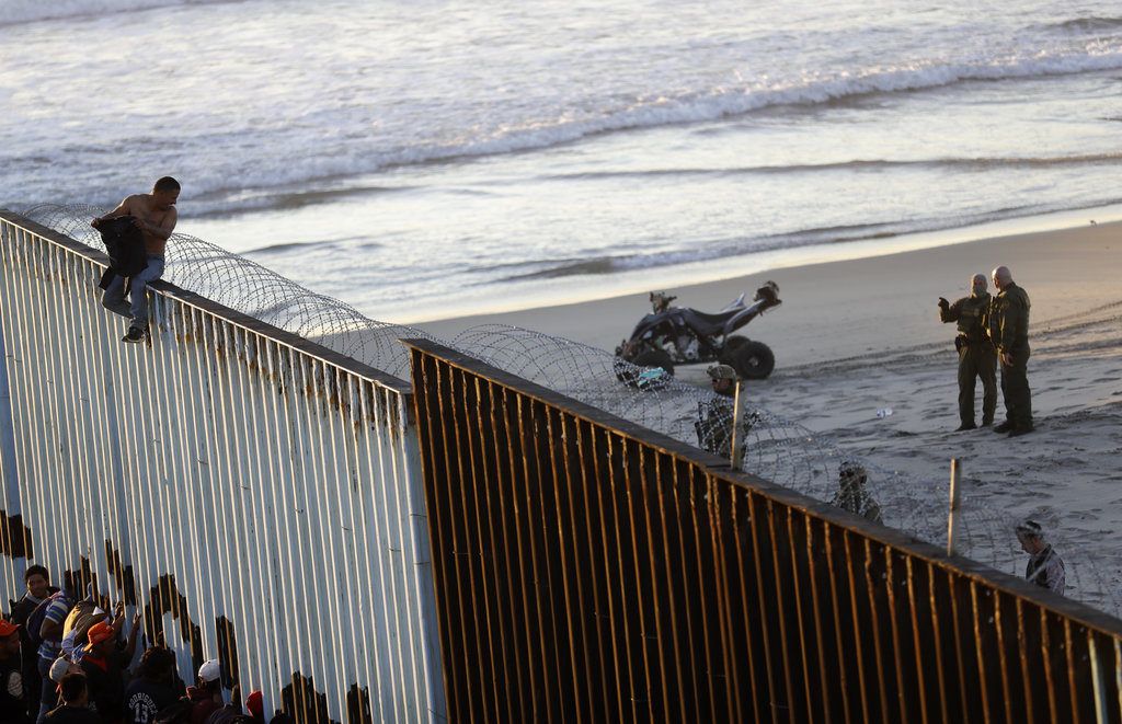Un migrante centroamericano está sentado sobre la estructura fronteriza que separa México (izquierda) de Estados Unidos (derecha), mientras agentes fronterizos estadounidenses vigilan la escena, el 14 de noviembre de 2018, en esta imagen tomada desde Tijuana, México. (AP Foto/Gregory Bull)