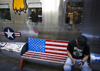 Un hombre navega en su smartphone en una banca decorada con la bandera estadounidense afuera de una tienda que vende marcas de Estados Unidos en un popular centro comercial en Beijing, el lunes 13 de mayo de 2019. (AP Foto/Andy Wong)