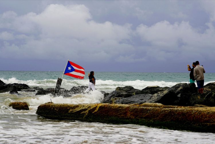 Una mujer posa para una fotografía con el mar y una bandera de Puerto Rico de fondo tras el paso de la tormenta tropical Dorian por la isla, en San Juan, Puerto Rico, el 28 de agosto de 2019. Foto: Ramón Espinosa/ AP.