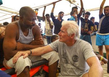 El actor Richard Gere, derecha, habla con migrantes al bordo del buque humanitario Open Arms durante el cruce del Mediterráneo, viernes 9 de agosto de 2019. (AP Foto/Valerio Nicolosi)
