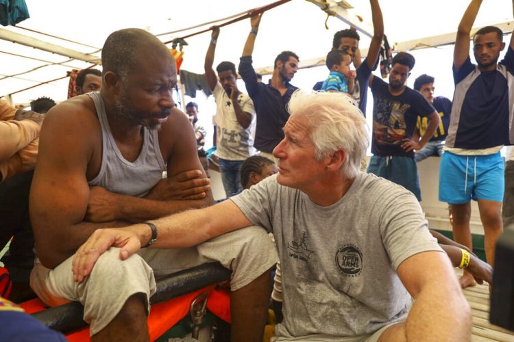 El actor Richard Gere, derecha, habla con migrantes al bordo del buque humanitario Open Arms durante el cruce del Mediterráneo, viernes 9 de agosto de 2019. (AP Foto/Valerio Nicolosi)