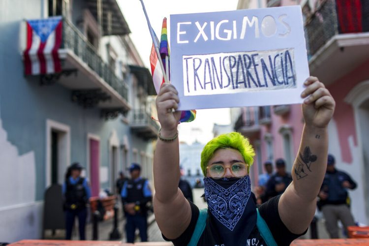 Un joven sostiene en lo alto un cartel durante una pequeña manifestación frente a La Fortaleza, la mansión del gobierno, en San Juan, Puerto Rico, el viernes 9 de agosto de 2019. (AP Foto/Dennis M. Rivera Pichardo)