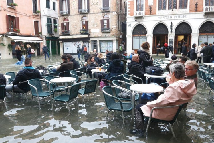 La vida continúa en medio de las inundaciones en Venecia y lugareños y turistas se sientan en un bar, en las mesas de la calle, a pesar de que el agua les sobrepasa los tobillos el 17 de noviembre del 2019. (Emiliano Creeps/ANSA via AP)