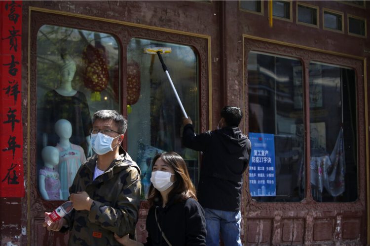 En esta imagen del 12 de abril de 2020, una pareja china camina frente a un trabajador que limpia las ventanas de una tienda de ropa donde se despliegan lineamientos para prevenir la pandemia en la reanudación de labores en Beijing. (AP Foto/Andy Wong)