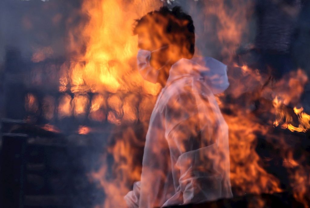 Un trabajador municipal con EPI visto tras las llamas de una de las hogueras prendidas en un crematorio para víctimas de la covid-19, este viernes en Bombay, India. Foto: EFE/ Divyakant Solanki.