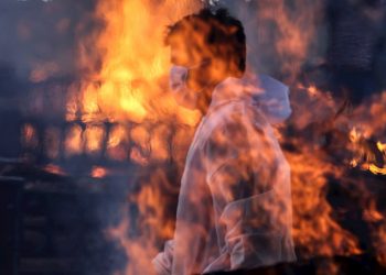 Un trabajador municipal con EPI visto tras las llamas de una de las hogueras prendidas en un crematorio para víctimas de la covid-19, este viernes en Bombay, India. Foto: EFE/ Divyakant Solanki.