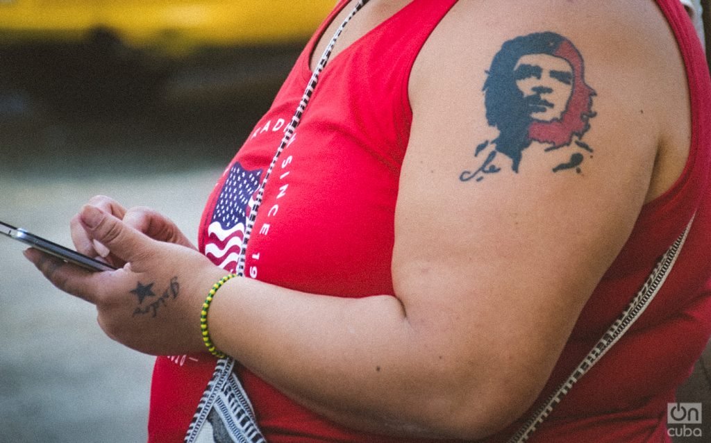 Detail of a Cuban woman at a Havana bus stop: Che Guevara is tattooed on her shoulder. On her wrist is an Orula bracelet, for the orisha of divination, the supreme oracle and the great benefactor of humanity and her chief advisor. She is wearing a t-shirt with the flag of the United States. Photo: Kaloian Santos