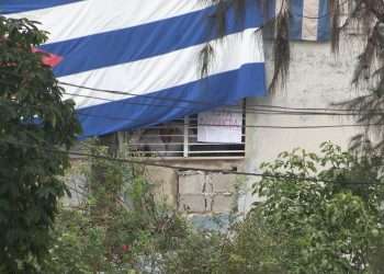El activista Yunior García Aguilera, uno de los convocantes de la marcha del 15 de noviembre, se asoma por su ventana con flores en la mano, mientras agentes vestidos de civil cubren su ventana con una bandera cubana. Foto tomada de la cuenta en Twitter del periodista Atahualpa Amerise.
