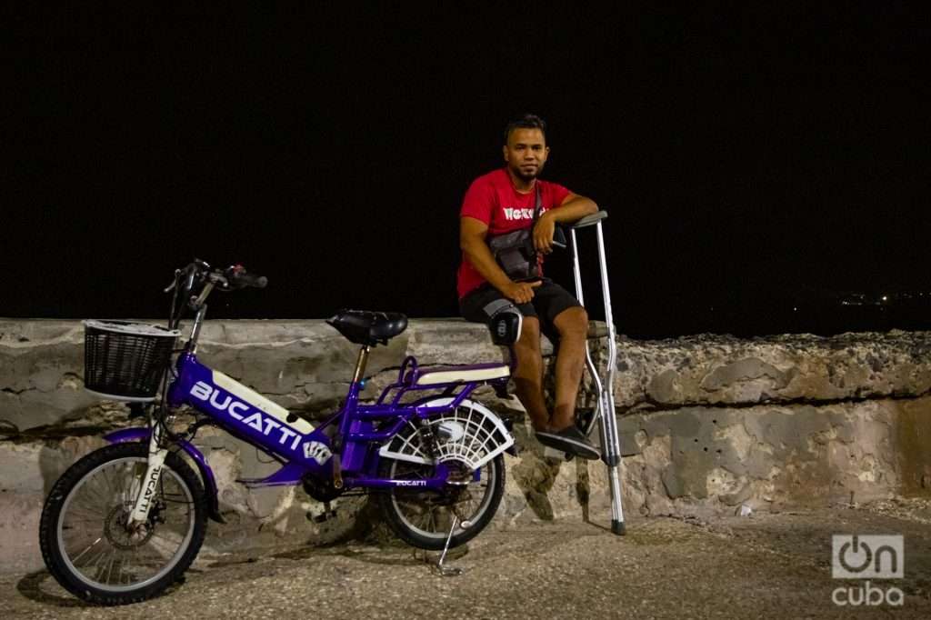 Hombre sentado de noche en el muro del malecón con su bicicleta eléctrica y una muleta, Habana, Cuba. Foto: Jorge Ricardo