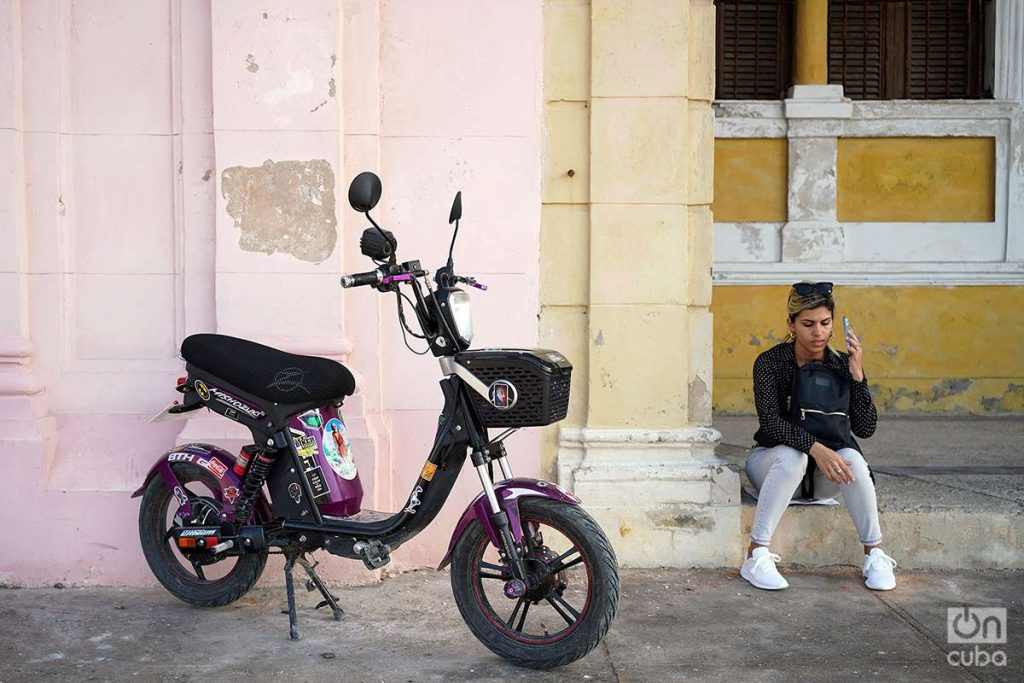 Mujer habla por celular junto a motocicleta eléctrica en La Habana, Cuba, 2023. Foto: Alejandro Ernesto.