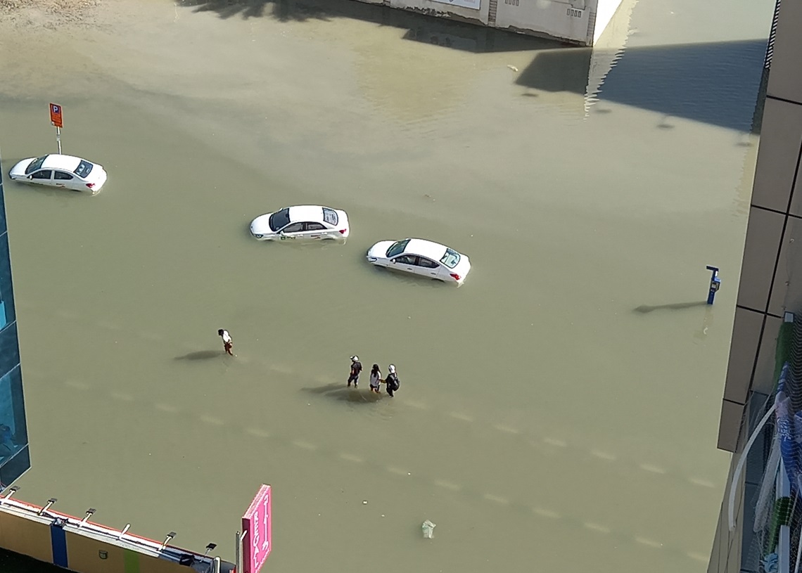 Foto desde mi balcón de carros abandonados y personas cruzando una calle inundada luego de las torrenciales lluvias en Dubái. 