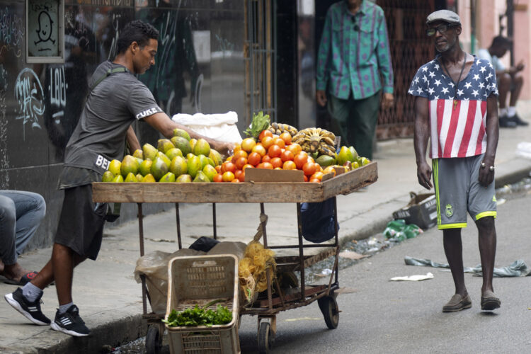 Un hombre con camiseta con la bandera de Estados Unidos pasa frente a un vendedor ambulante de verduras, en La Habana. Foto:  Yander Zamora/EFE.