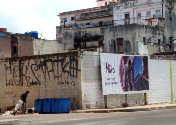 Un hombre registra un vertedero de basura a metros de una propaganda gubernamental sobre el 1 de mayo, en La Habana. Foto: AMD.