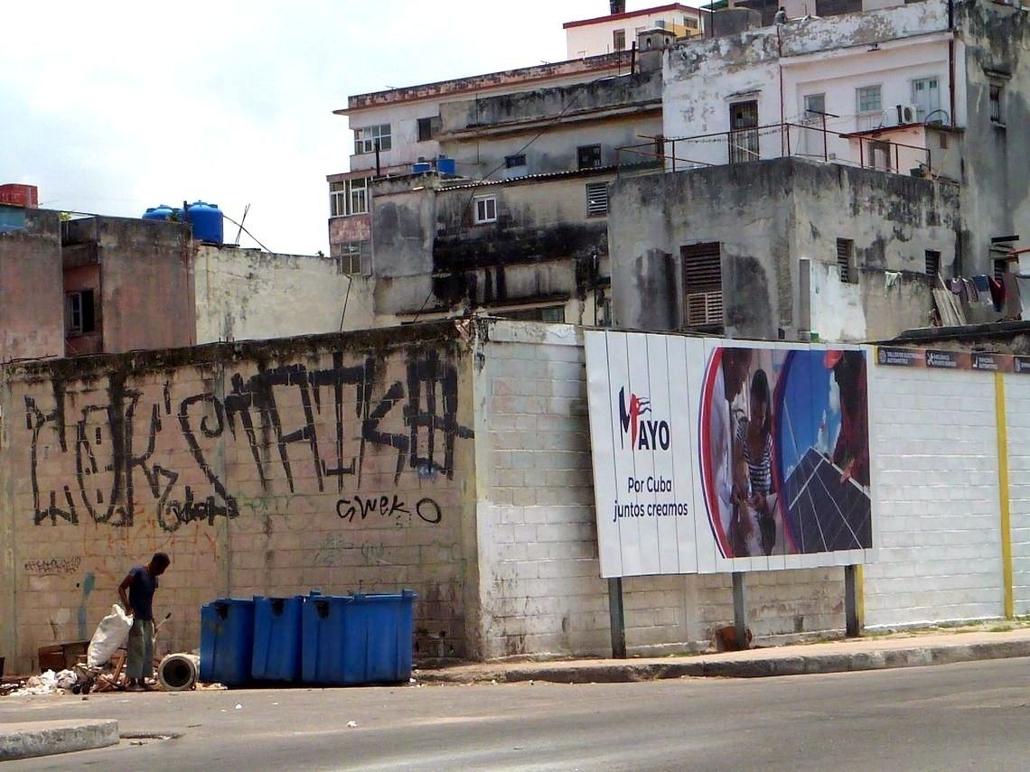 Un hombre registra un vertedero de basura a metros de una propaganda gubernamental sobre el 1 de mayo, en La Habana. Foto: AMD.