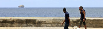 Dos hombres caminan por el Malecón de La Habana, mientras un barco se aleja de Cuba. Foto: Ernesto Mastrascusa / EFE.