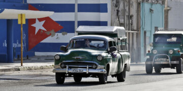 Vehículos antiguos transitan frente a un mural con la bandera de Cuba pintada. Foto: EFE/Ernesto Mastrascusa.